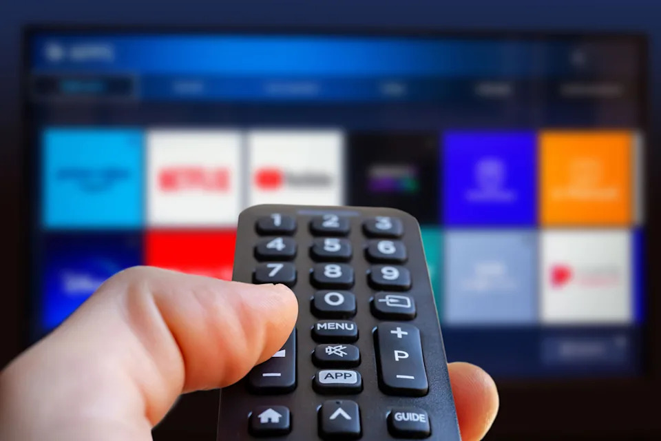 A person using a remote control in front of a TV showing app icons related to streaming services