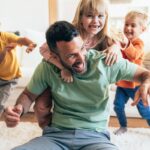 A happy single father and his three children are playing on the floor in the living room at home. A loving father spends free time with his children. They enjoy the weekend at home. Three children are playing with their father on the floor.