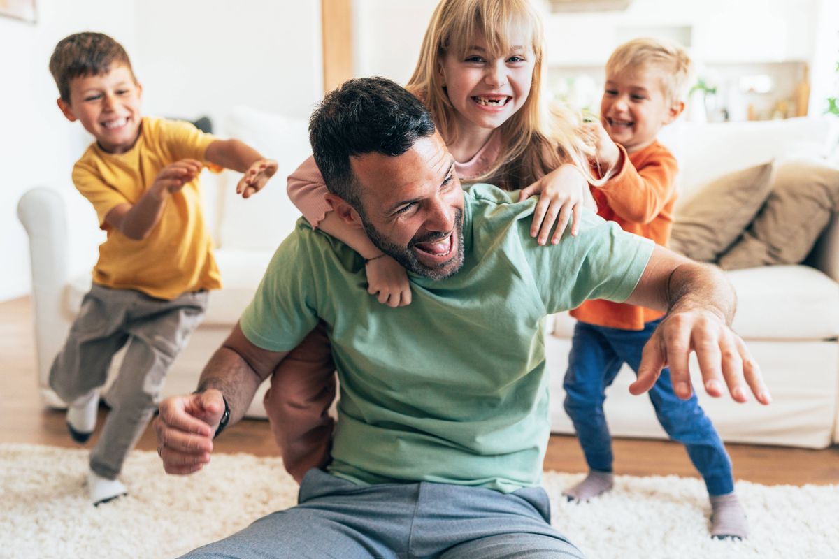 A happy single father and his three children are playing on the floor in the living room at home. A loving father spends free time with his children. They enjoy the weekend at home. Three children are playing with their father on the floor.