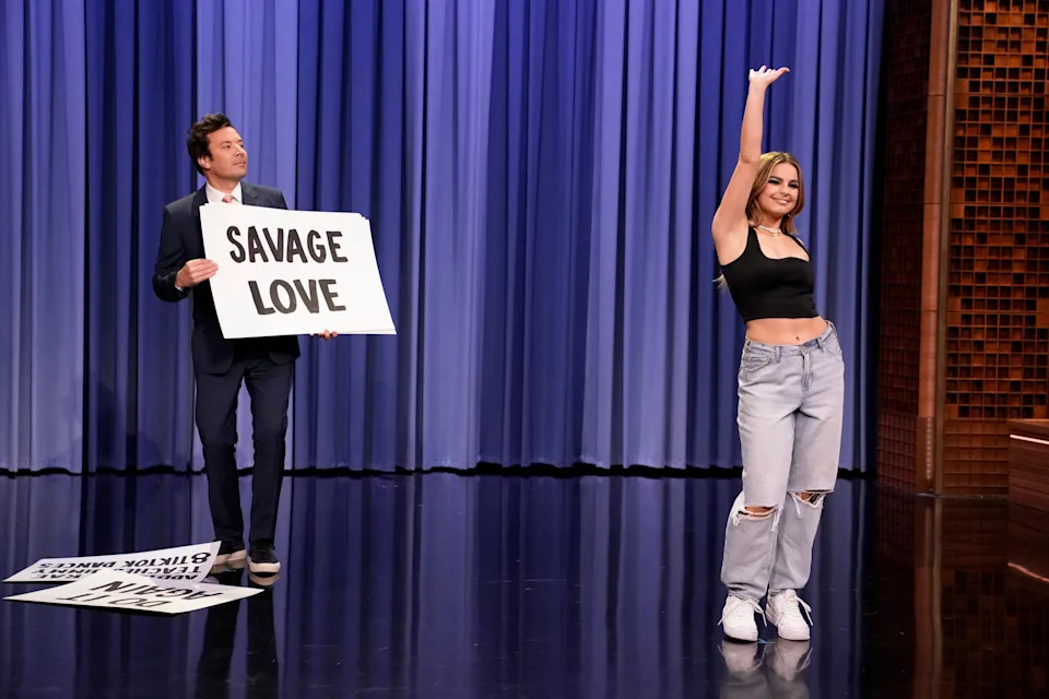 The TV host is holding a "Cruel Love" sign as a woman in a crop top and ripped jeans rock the stage