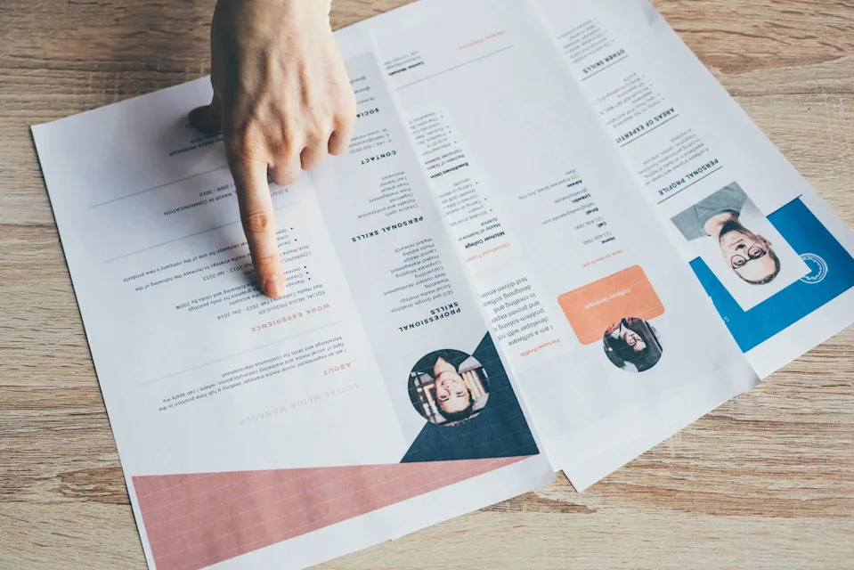 A hand points to a text on a list of copies printed on a wooden table, highlighting different parts and photos of different people.